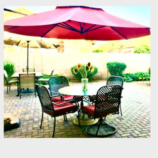 A cozy patio setup featuring a round table with red cushioned chairs under a large red umbrella, surrounded by a brick floor and garden plants.