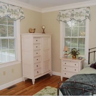A cozy bedroom featuring a tall dresser, a nightstand with decor, and large windows with floral curtains.