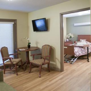 A healthcare facility room featuring a medical bed, seating area with chairs and table, and a wall-mounted TV.