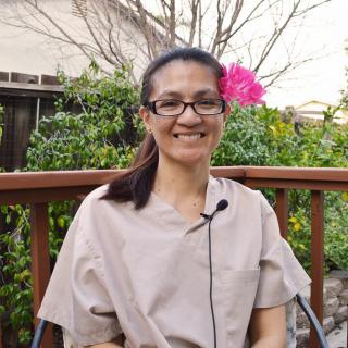 A woman with glasses is sitting outdoors, smiling and wearing a microphone, with a pink flower behind her.