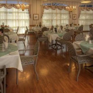 An elegant dining area with neatly arranged tables and chairs, featuring large windows with curtains and wooden flooring.