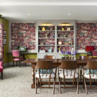 A vibrant dining room featuring a large wooden table surrounded by chairs, with colorful wallpaper and bookshelves in the background.