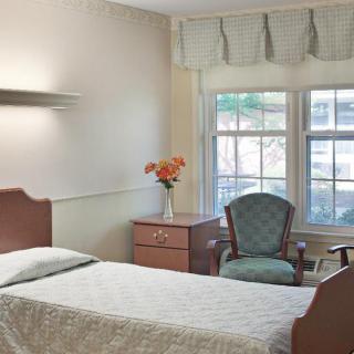 A simple hospital room with a single bed, nightstand, chair, and flowers by the window.