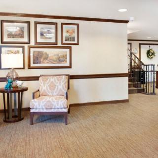 A cozy interior space featuring a patterned chair, side table with a lamp, framed artwork on the wall, and a staircase in the background.