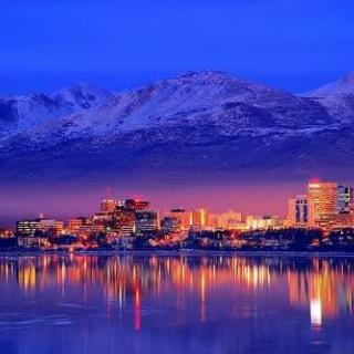 A stunning cityscape at dusk, showcasing the reflection of illuminated buildings against a serene body of water, with majestic mountains in the background.