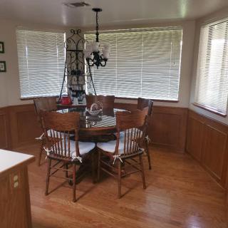 A cozy dining room with wooden flooring and a round table surrounded by chairs, illuminated by a chandelier, with windows featuring blinds.