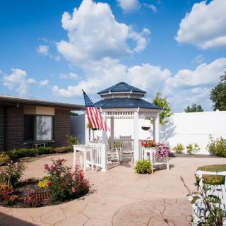 A peaceful courtyard featuring a gazebo, American flag, and vibrant plant life under a clear blue sky.