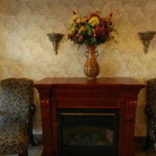 A cozy living room setup featuring a wooden fireplace adorned with a vase of colorful flowers, flanked by two patterned chairs and wall sconces.