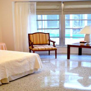 A spacious bedroom with a bed, armchair, and table next to large windows.