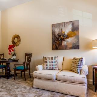 A cozy living room with a beige sofa, decorative pillows, and abstract artwork on the wall. A small table and chairs are tucked in the corner.