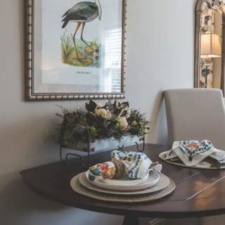 A cozy dining room with a neatly set table, featuring decorative plates and a floral centerpiece. A framed bird illustration hangs on the wall, adding to the room's charm.