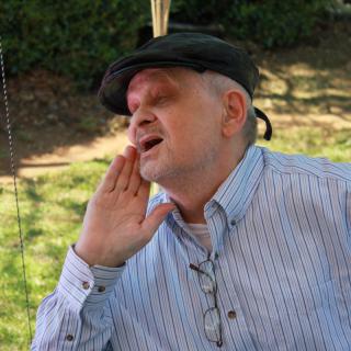 A man wearing a hat and glasses holds his hand to his mouth while outdoors.