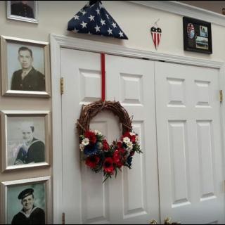 A wall decorated with framed military photographs, an American flag folded in a triangle, and a wreath hanging on double doors.