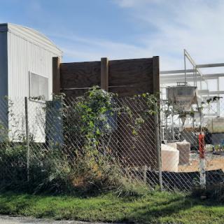 A view of an industrial site surrounded by a chain-link fence and overgrown foliage.