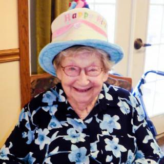 An elderly woman wearing a festive birthday hat and a floral blouse, smiling indoors.