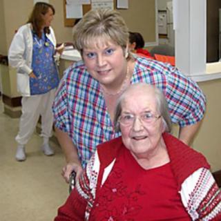 An elderly woman in a red outfit is smiling while a healthcare worker stands beside her.