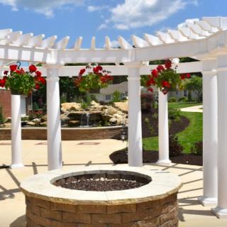 A beautifully landscaped garden with white pergola and hanging flowers surrounding a fire pit.