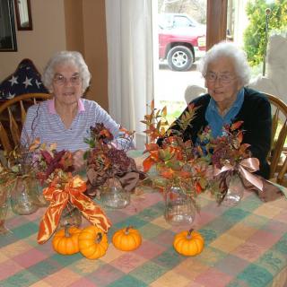 Two elderly women sitting at a table decorated with autumn-themed flowers and small pumpkins.
