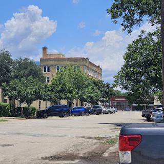 A view of a historic building surrounded by trees and parked vehicles under a partly cloudy sky.