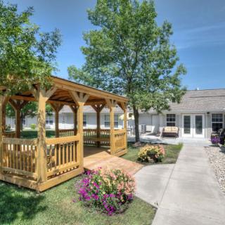 A sunny garden scene featuring a wooden gazebo, surrounded by trees and flowers with a pathway leading to a building.