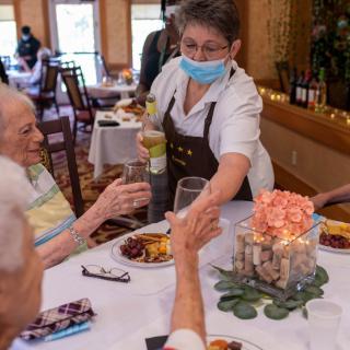 A waitress wearing a mask serves drinks to seniors at a restaurant table.