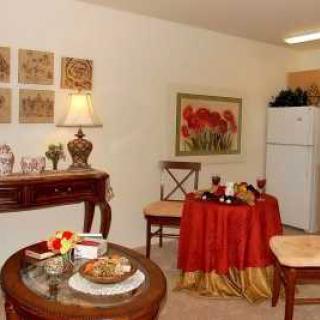 A cozy dining and living area with classic decor, featuring a small table draped in red cloth, a lamp on a side console, and a refrigerator in the background.