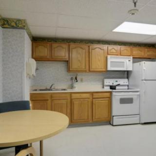 A simple kitchen setup with wooden cabinets, a stove, microwave, refrigerator, and a small round table with chairs.