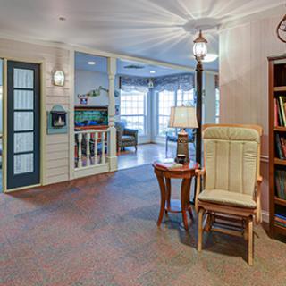 A cozy living room with a rocking chair, lamp, and bookshelf near large windows.