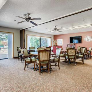 A spacious dining room with multiple tables and chairs, featuring ceiling fans, large windows, and a television on the wall.
