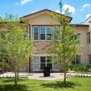 A two-story residential house with a stone facade, surrounded by trees and a green lawn under a clear blue sky.