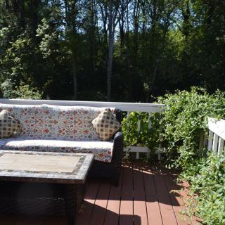 A cozy outdoor patio with a sofa, cushions, and a table set on a wooden deck surrounded by greenery.