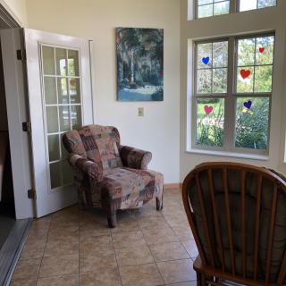 A cozy living room corner with a patterned armchair, art on the wall, and sunlight streaming through decorated windows.