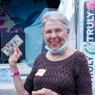 A smiling woman holding a bill outdoors, with a mask partially down.