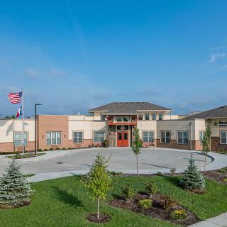 A modern school building with a circular driveway, landscaped grounds, and a flagpole against a clear blue sky.