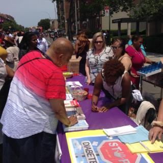 A lively community event with people engaging at an information booth under the summer sun.