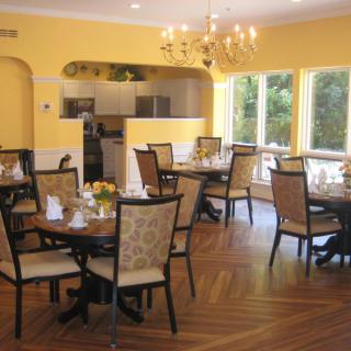 A cozy dining area with elegantly set tables and chairs, illuminated by a chandelier and abundant natural light.