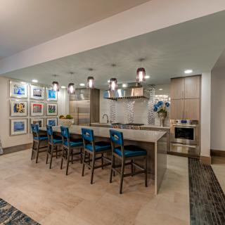 A modern kitchen with a large island, blue bar stools, and pendant lighting, featuring artwork on the walls.