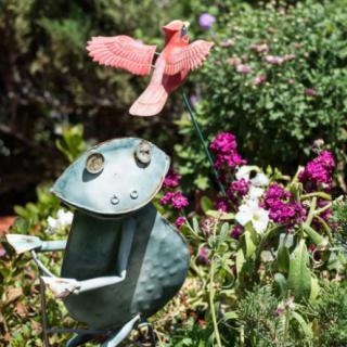 A whimsical garden scene featuring a metal frog sculpture among vibrant flowers, with a red metal bird flying overhead.