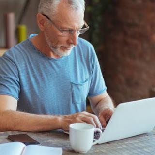 A man in a blue t-shirt working on a laptop at a table with a coffee cup.