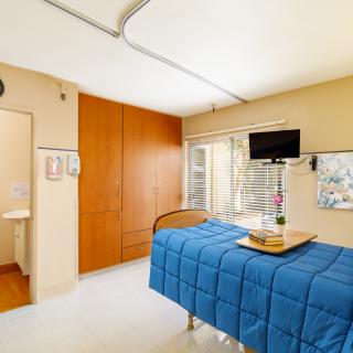 A clean and bright hospital room featuring a single bed with blue bedding, a wall-mounted television, and a vase of flowers by the window.