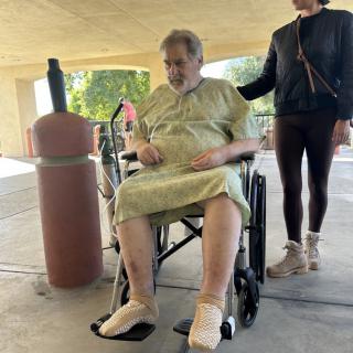 An elderly person in a wheelchair wearing a hospital gown, accompanied by a caregiver outdoors.