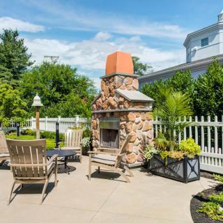 A sunny outdoor patio featuring a stone fireplace, surrounded by chairs and lush greenery.