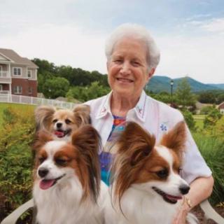 An elderly woman smiling with three dogs in an outdoor garden setting.