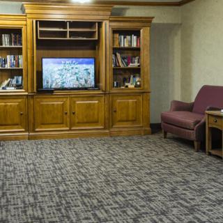 A cozy living room featuring a wooden bookshelf and cabinet combination, with a TV at the center, surrounded by books and a maroon chair on carpeted flooring.
