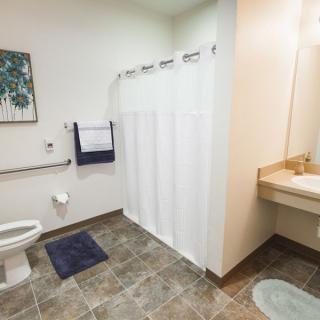A spacious and accessible bathroom with a tiled floor, featuring a shower with a curtain, grab bars, a toilet, and a sink.