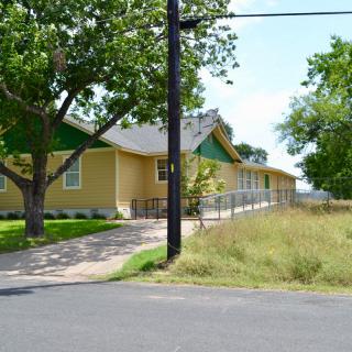 A single-story yellow house with a sloped roof, surrounded by trees and grass, next to a paved pathway.