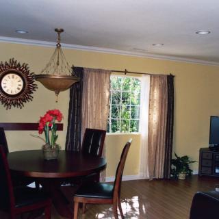 A bright living and dining area with a wooden table and chairs, a decorative clock on the wall, and sunlight streaming through the window.