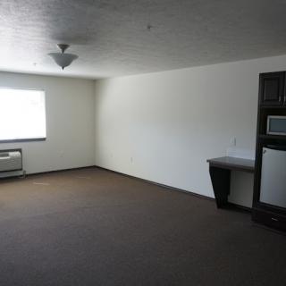 An empty apartment room with a window, air conditioner, and small kitchenette.