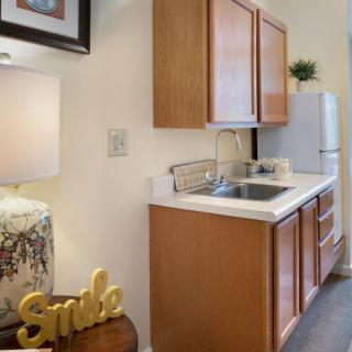 A compact kitchen area with wooden cabinets, a sink, and a refrigerator, accented by a decorative lamp and 'Smile' sign.