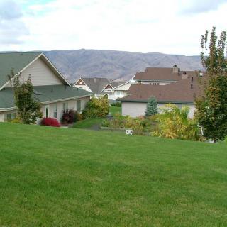 A suburban neighborhood with green lawns and houses set against a backdrop of mountains under a cloudy sky.